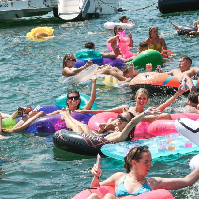 A group of people is enjoying a sunny day, floating on colorful inflatable tubes in a body of water. They are smiling, waving, and wearing swimwear. There's a festive atmosphere, with a snowman float and someone wearing a Santa hat.