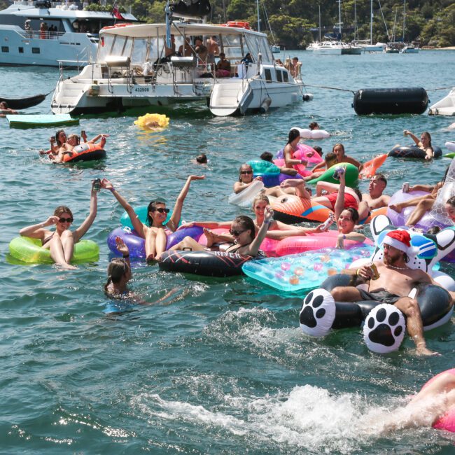 A lively scene of people enjoying a sunny day on the water, lounging on colorful inflatable floats and swimming. Boats are anchored in the background. The water is clear and the atmosphere is festive.