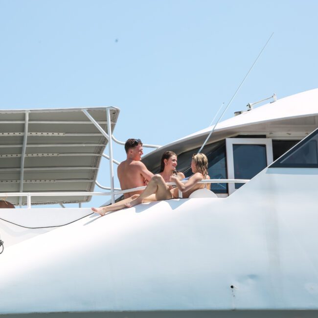 Three people are sitting and relaxing on the deck of a white yacht, enjoying sunny weather. The yacht is anchored, and a clear blue sky forms the backdrop.