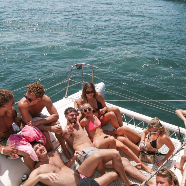 A group of people in swimwear relax on a catamaran's net over the ocean. They're smiling and lounging, with some holding drinks, enjoying a sunny day on the water. The sea is calm with a few ripples.