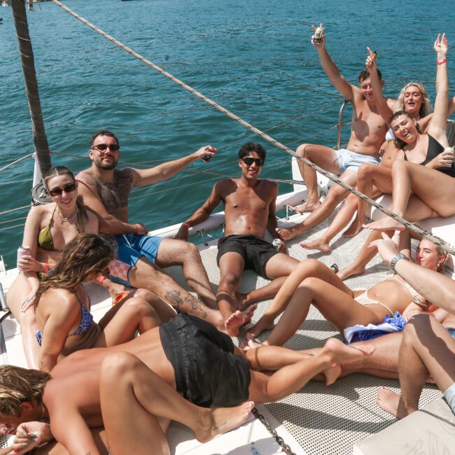 A group of people in swimsuits are relaxing and cheering on a catamaran deck surrounded by water. They appear to be enjoying drinks and sunbathing under a clear sky. A yacht is visible in the background.