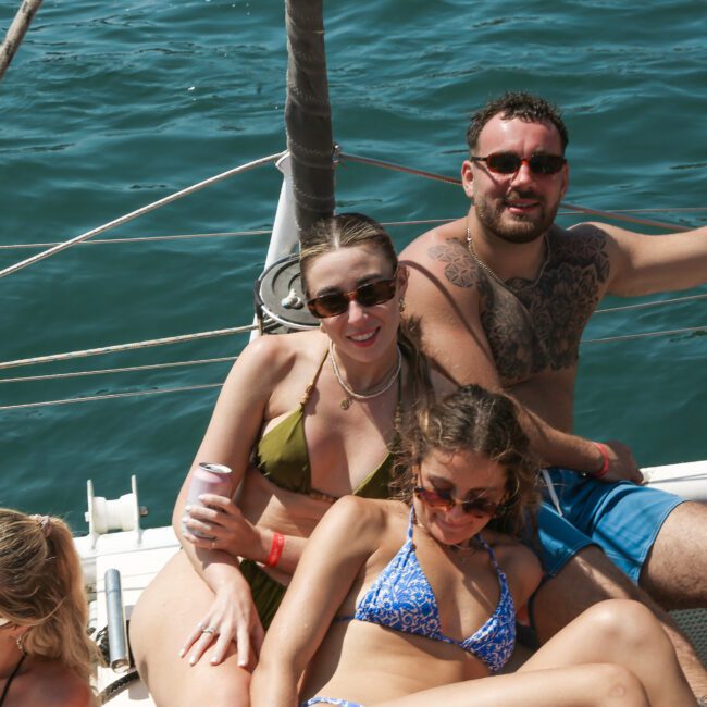 Four people are relaxing on a boat, enjoying a sunny day. Two women are in bikinis, and a man in shorts is beside them. They appear to be on a catamaran with blue water in the background. Everyone seems relaxed and happy.