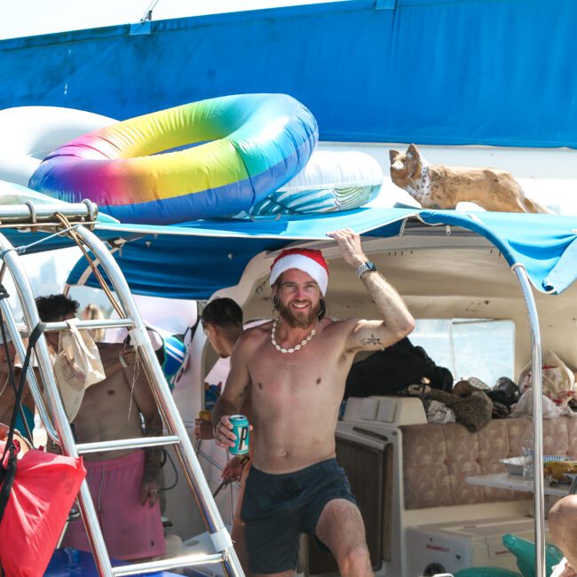 A group of people on a boat enjoying a sunny day. They're wearing Santa hats, and one person is holding a drink, smiling. There are colorful inflatables and a dog resting on the top of the cabin. The scene is festive and lively.