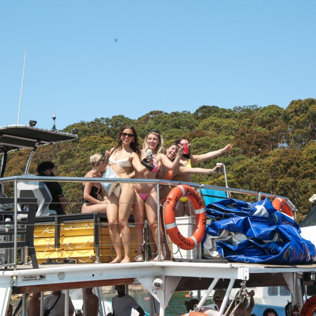 A group of people on a boat, with two women in bikinis posing and smiling on the upper deck. The surrounding scenery includes trees and another boat in the background. The weather appears sunny and clear.