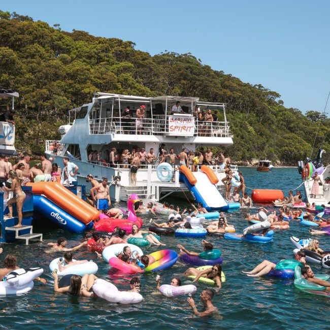 A lively scene with a group of people enjoying a sunny day on a lake. They are floating on colorful inflatables and swimming near several boats. The dense green foliage of the shoreline provides a vibrant natural backdrop.