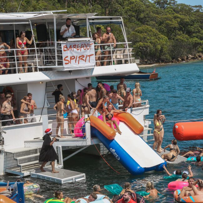 A large group of people enjoy a party on a boat named "Barefoot Spirit." Some people are on the deck, while others swim in the water with inflatable toys. The scene is lively, set against a backdrop of water and trees.