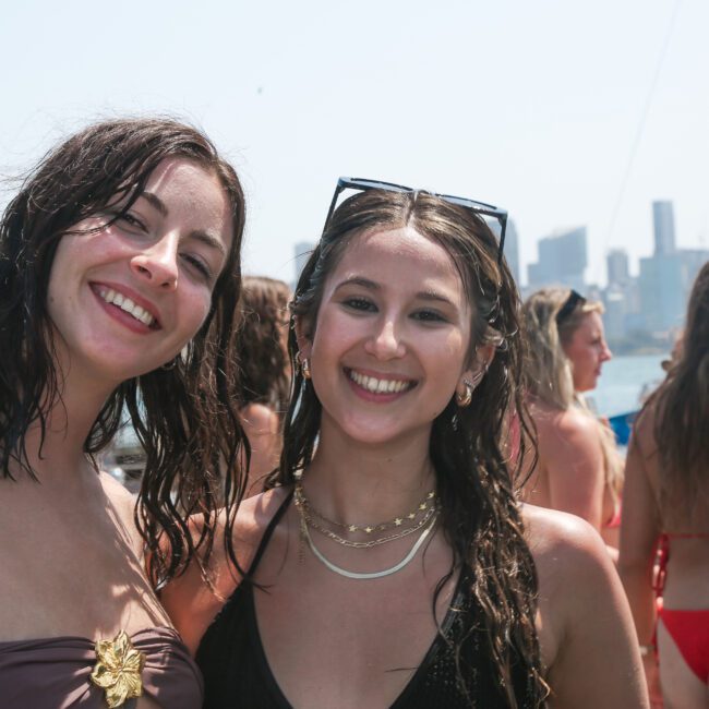 Two smiling women with wet hair are standing outdoors on a sunny day, posing for a photo. They are wearing swimsuits and have jewelry on. In the background, several other people are enjoying a day near the water with boats and a city skyline visible.