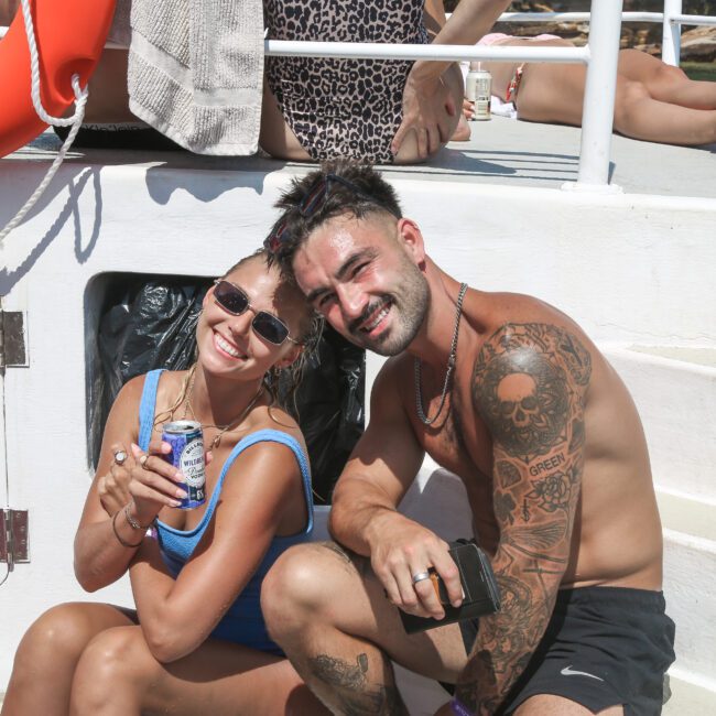 A woman in a blue swimsuit and sunglasses holds a drink can, smiling beside a shirtless man with tattoos who is also smiling. They are sitting on a boat with other people relaxing in the background and life rings nearby.