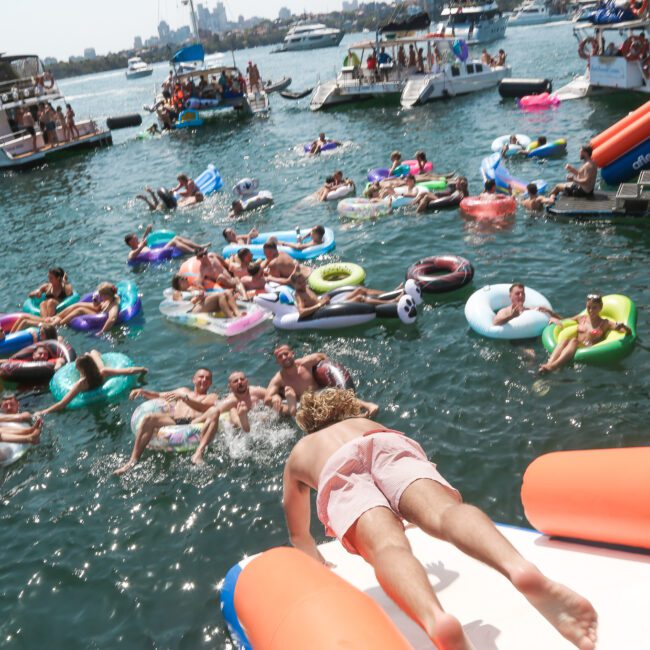 A person slides into the water on an inflatable water slide as a large group of people on colorful inflatables relax and enjoy the sunny day. Boats are in the background on the lively, crowded waterfront.