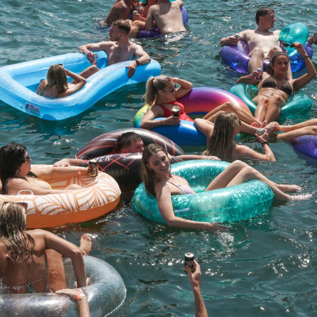 A group of people floating on colorful inflatable tubes and rafts in clear water, enjoying a sunny day. Some are holding drinks, and they all appear relaxed and having fun.