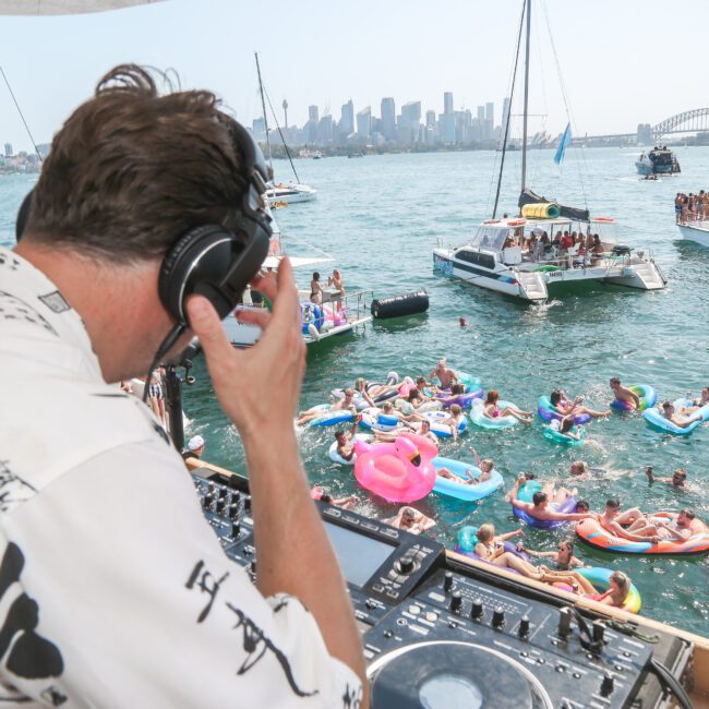 A DJ wearing headphones performs on a boat while a crowd of people on colorful inflatables enjoy a party in the water. The Sydney skyline and the Harbour Bridge are visible in the background under a clear blue sky.