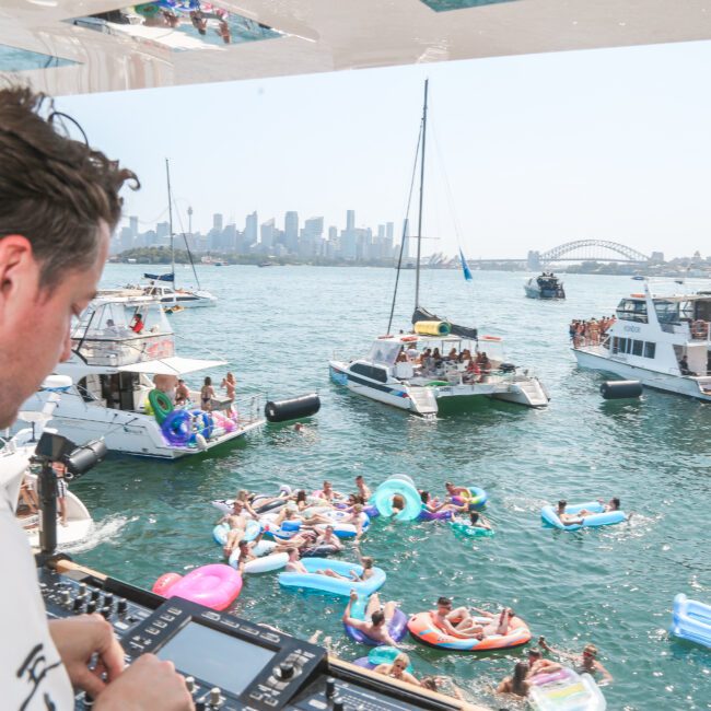 A DJ performs on a boat overlooking a lively party scene on the water. People relax on inflatables and nearby boats, with a city skyline and bridge visible in the background under a clear blue sky.