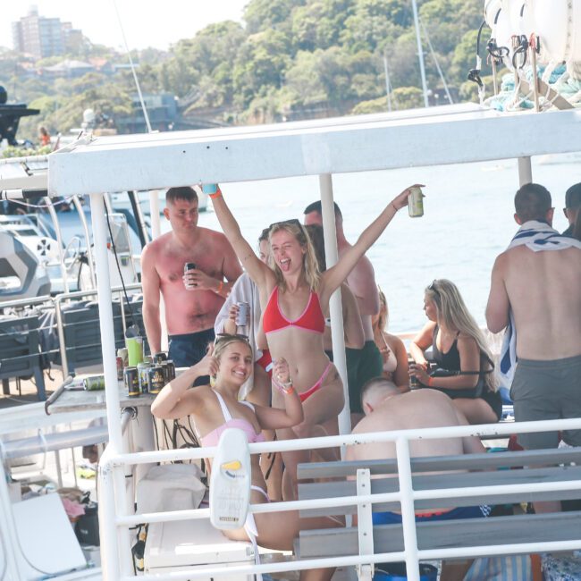 A group of people in swimwear enjoying a boat party. Some individuals are sitting and others are standing, holding drinks. The background features water, trees, and other boats, creating a lively and festive atmosphere.