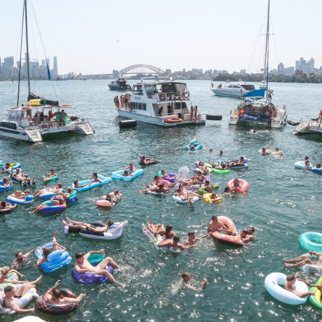 A lively harbor scene with numerous people relaxing on colorful inflatables in the water. Several boats are anchored nearby. The skyline and iconic bridge are visible in the background, indicating a festive, sunny day.