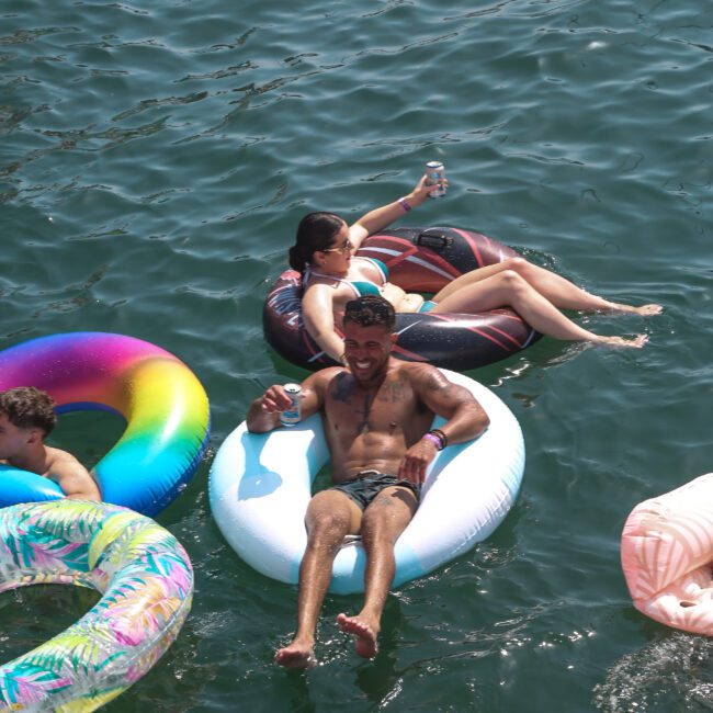 People are floating on inflatable tubes in a body of water. One person is holding a drink and smiling, while others relax on colorful inflatables around them. The scene is lively and sunny.