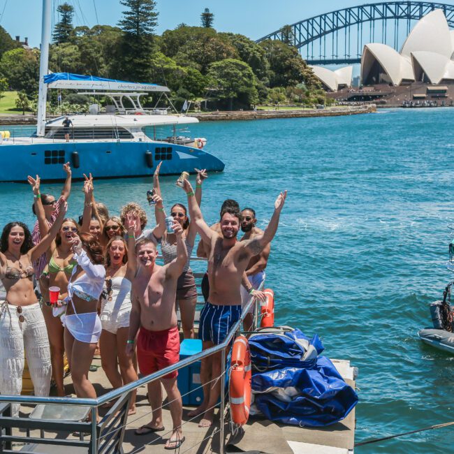 A group of people on a boat posing happily with arms raised. They are near a body of water with the Sydney Opera House and Sydney Harbour Bridge in the background. Other boats are visible on the water under a sunny sky.