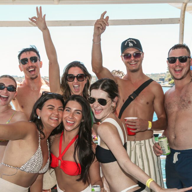 A group of smiling people in swimsuits enjoying a sunny day on a boat. They are posing with their arms raised, drinks in hand. The ocean is visible in the background.