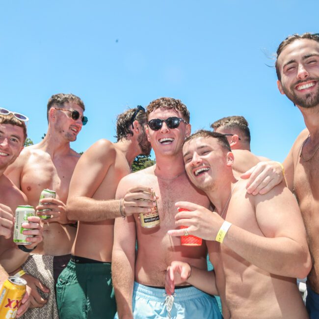 A group of shirtless young men smiling and enjoying a sunny day together. They are holding cans and seem to be in a festive and relaxed mood against a clear blue sky.