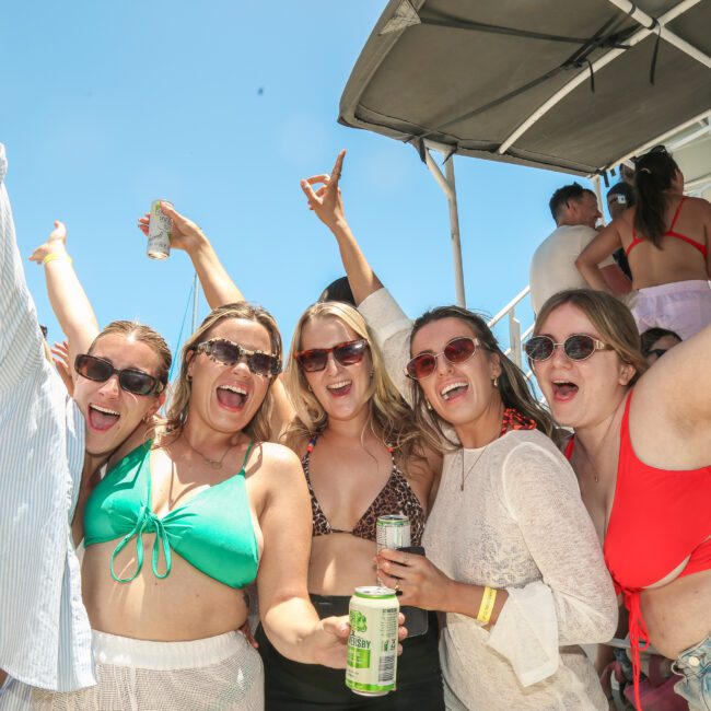 A group of five people smiling and posing on a boat under a clear blue sky. They are wearing sunglasses and summer attire, holding drinks, and appear to be enjoying a festive atmosphere with others in the background.