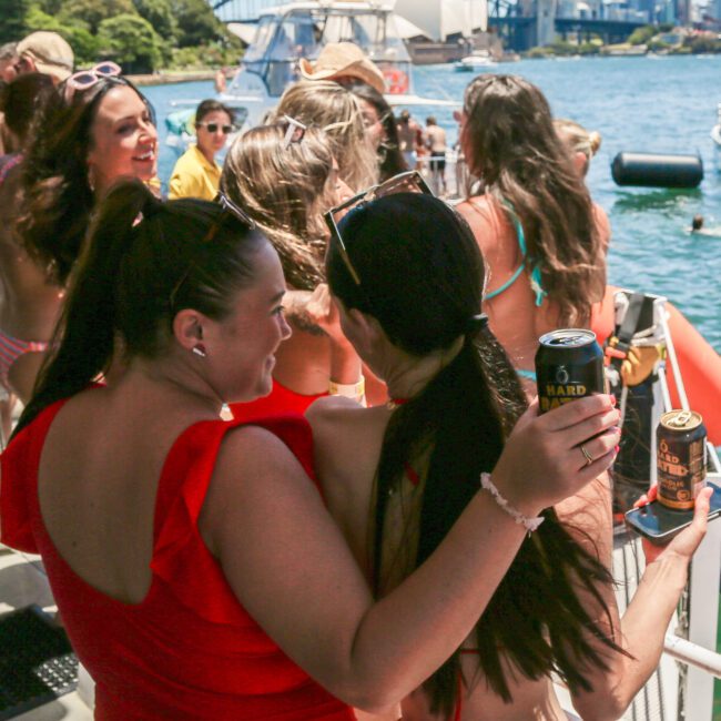 A group of people is enjoying a sunny day on a boat, with the Sydney Opera House and Harbour Bridge in the background. Two women in vibrant outfits are in the foreground, holding canned drinks and looking at the scenic view.