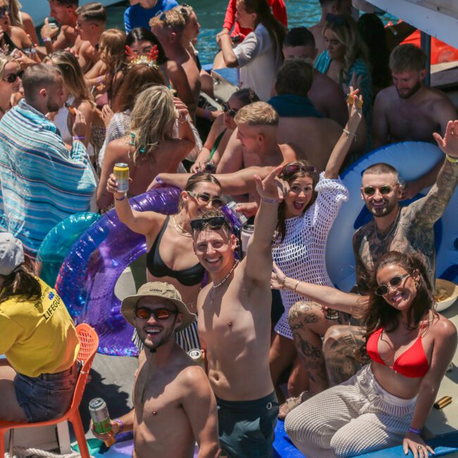 A large group of people are enjoying a lively boat party. Many are smiling, waving, and wearing swimwear. Inflatable floats and drinks are visible. The sun is shining, creating a vibrant and festive atmosphere.