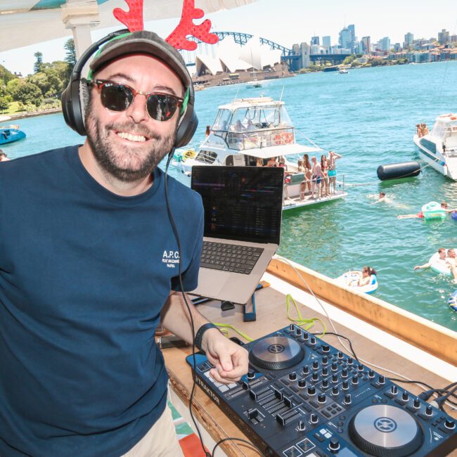 A man wearing reindeer antlers and sunglasses is smiling at a DJ booth on a boat. In the background, people are enjoying a sunny day on inflatables with a view of the Sydney Opera House and other boats on the water.