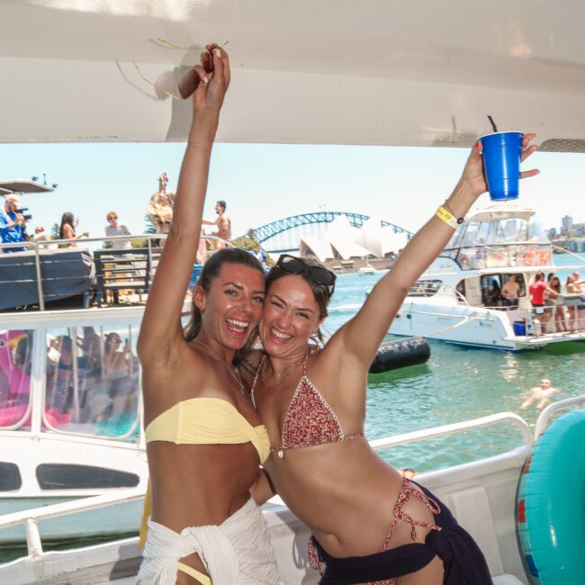 Two women in swimsuits smile and pose on a boat at a lively outdoor event. One wears a yellow bikini with a towel wrapped around her waist, and the other wears a patterned bikini. Boats and people are visible in the background under a sunny sky.