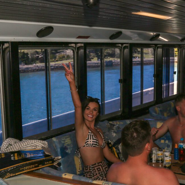 A woman in a patterned swimsuit is sitting inside a boat, smiling and flashing a peace sign. She is surrounded by other people and the boat's windows show a view of water and land outside. Cans and items are on the table in front of her.