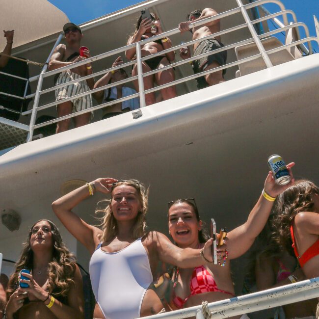 A group of people are enjoying a sunny day on a boat. Some are holding drinks and smiling, while others are waving. The atmosphere is lively, and the sky above is clear and blue.