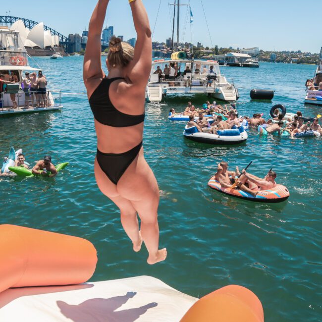 A person in a black bikini jumps from a boat into a harbor filled with people on inflatables. Several boats are anchored nearby, and a bridge is visible in the background under a clear blue sky.