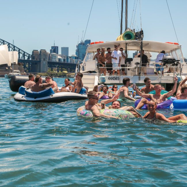 A group of people relax on inflatables in the water near a boat. The Sydney Harbour Bridge and Sydney Opera House are visible in the background under a clear blue sky.