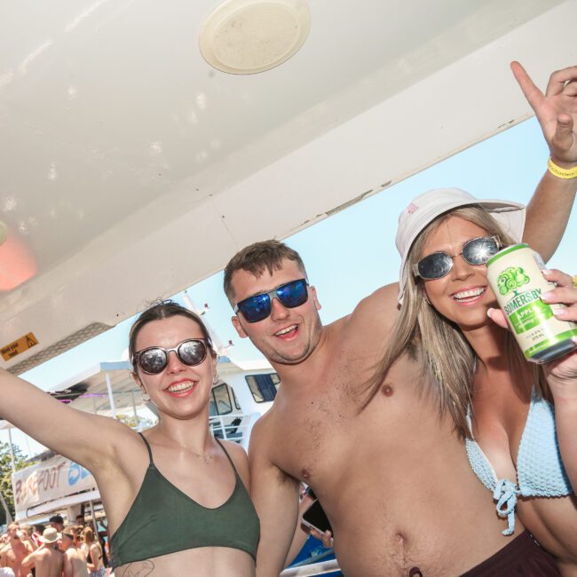 Three people in swimsuits and sunglasses enjoying a party on a boat. The person on the right holds a green drink can. They're smiling and have their arms around each other, with others in the background. Trees and blue sky are visible.