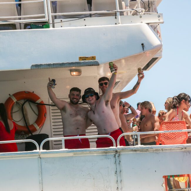 A group of people enjoying a sunny day on a boat. Three men at the railing are smiling and making hand gestures. The deck is crowded with others, some wearing swimsuits and hats. One person in the foreground holds a bag.