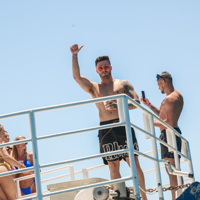 A man in black shorts and orange sunglasses stands on a boat deck, raising one hand. Another man is taking a photo with his phone. Two women in swimsuits relax nearby against a blue sky.