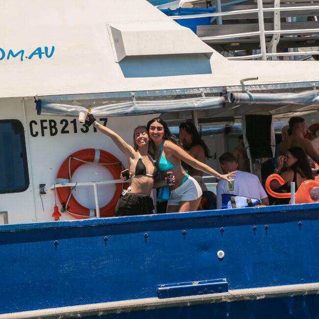 Two people stand on a boat, smiling and taking a selfie. It's a sunny day, and others are relaxing on the boat. A lifebuoy is visible on the side, and the boat is painted white and blue.