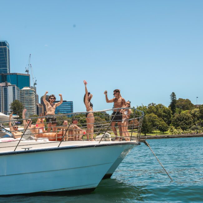 A group of people are enjoying themselves on a yacht in a sunny harbor. Some are standing and waving, with city skyscrapers and greenery in the background. The bright blue sky and clear water create a vibrant atmosphere.