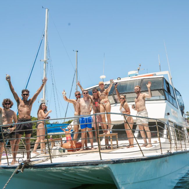 A group of people in swimwear celebrate and pose on the deck of a yacht under a clear blue sky. They are waving and holding drinks, enjoying a sunny day on the water. Two other boats are in the background with more people.