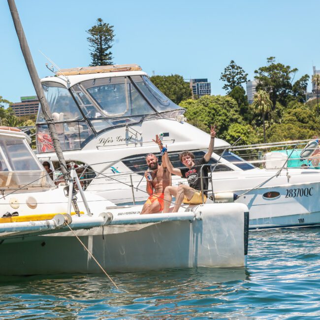 A group of people on a sailboat and nearby yachts are enjoying a sunny day on the water. The city skyline is visible in the background. Two individuals on the sailboat are raising drinks and smiling at the camera.