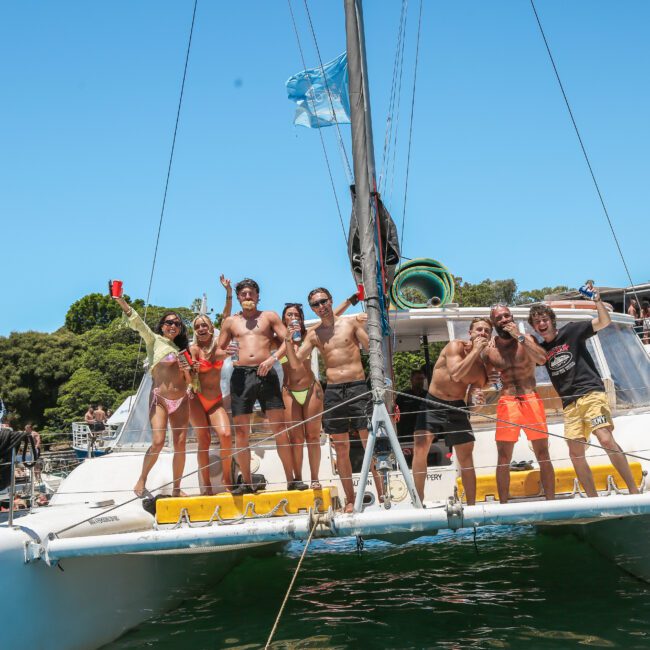 A group of people stand on the deck of a catamaran, posing and smiling in swimwear under a clear blue sky. They are docked in a harbor with other boats and lush greenery in the background.