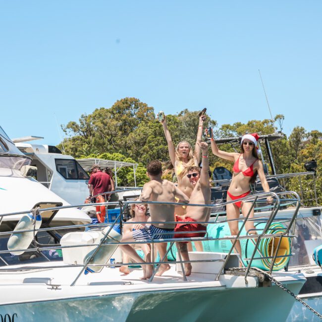 A group of people enjoying a sunny day on a boat. Some wear swimsuits, and one person is in a Santa hat, raising an arm in celebration. Trees and a clear blue sky are in the background.