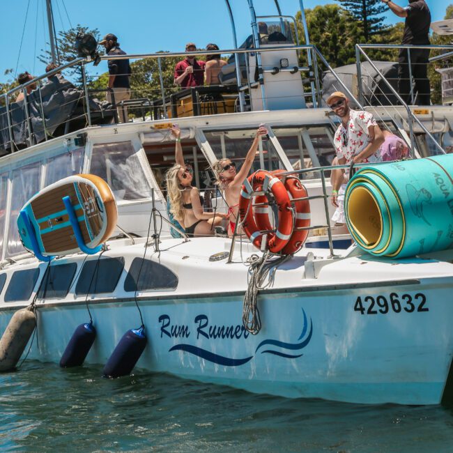 A group of people are enjoying a sunny day on a boat named "Rum Runner." They are smiling and posing for the camera. The boat is docked, surrounded by water and nearby vessels. In the background, trees and people on another boat can be seen.