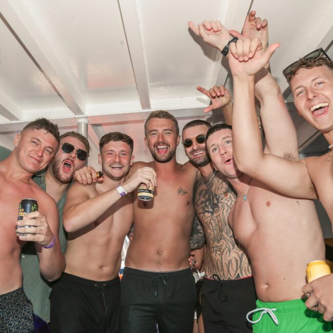 A group of shirtless men is celebrating on a boat. Several are holding drinks and smiling at the camera. They have raised arms, some making hand gestures, and appear to be having a fun time.