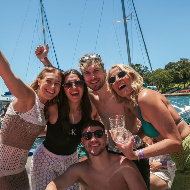 A group of five people smiling and posing on a boat under a clear blue sky. They are wearing casual summer clothing, and the background features sailboats and lush greenery along the shoreline.