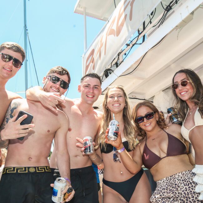 A group of six adults smiling and posing on a boat under a sunny sky. They are wearing swimwear and holding canned drinks, enjoying a lively and upbeat atmosphere.