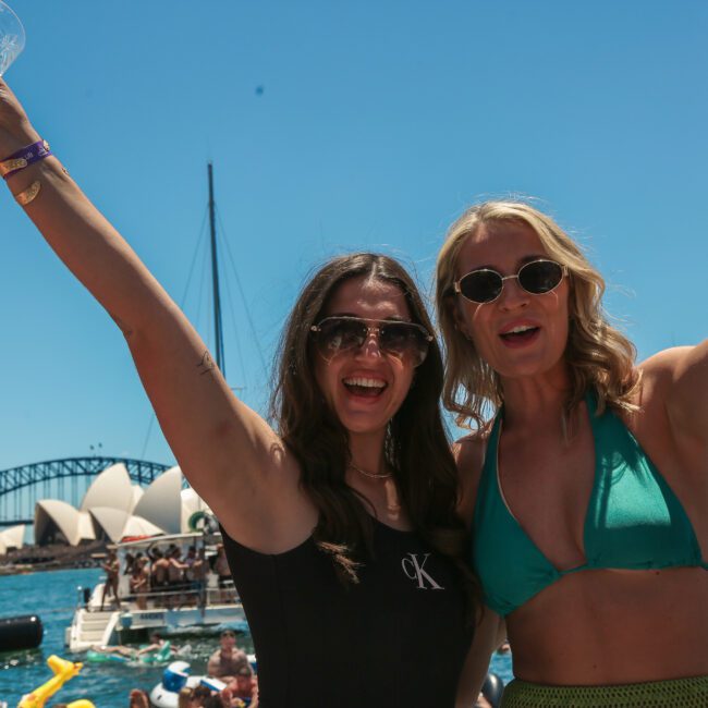 Two women in swimsuits cheerfully raise their arms, holding wine glasses, against a backdrop of the Sydney Opera House and Harbour Bridge. The scene is lively, with a boat and other people enjoying the sunny day on the water.