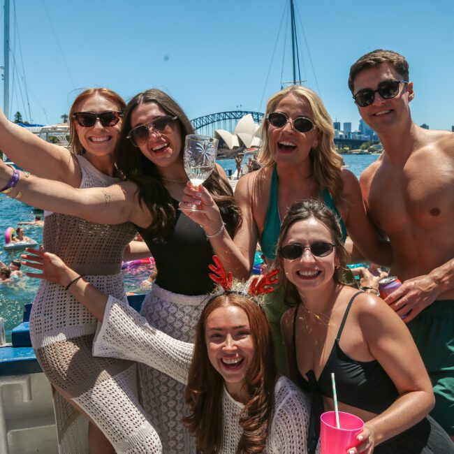 A group of people on a boat celebrating, with the Sydney Opera House and Harbour Bridge in the background. They are smiling, holding drinks, and appear to be enjoying a sunny day. The water is filled with people swimming and having fun.