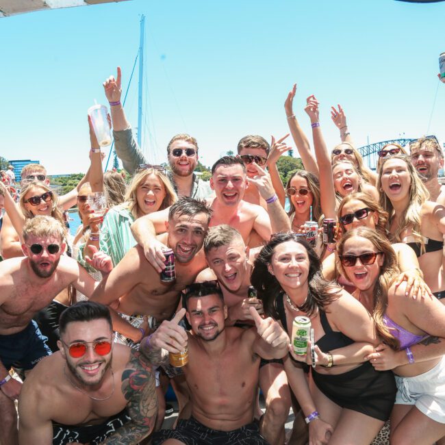 A large group of people are celebrating on a boat under a sunny sky. They are smiling, holding drinks, and wearing summer attire. The background features a city skyline and a bridge.