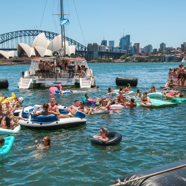 People enjoy a sunny day floating on inflatables in a harbor. The Sydney Opera House and Harbour Bridge are visible in the background. Boats are docked around, with more people relaxing on board.