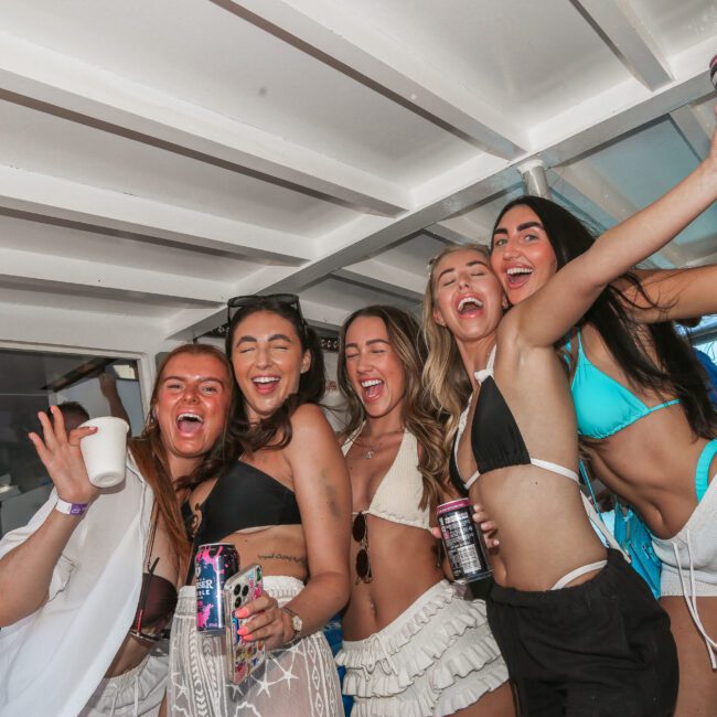 A group of five women smiling and posing together on a boat. They are wearing swimsuits and holding cans, with a lively and joyful atmosphere. The city skyline and greenery can be seen in the background.