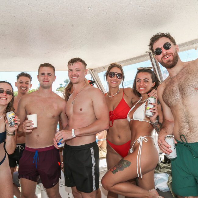 A group of people in swimwear smile and pose for a photo inside a boat. They are holding drinks and appear to be enjoying a sunny day, with a partial view of the water and city skyline in the background.